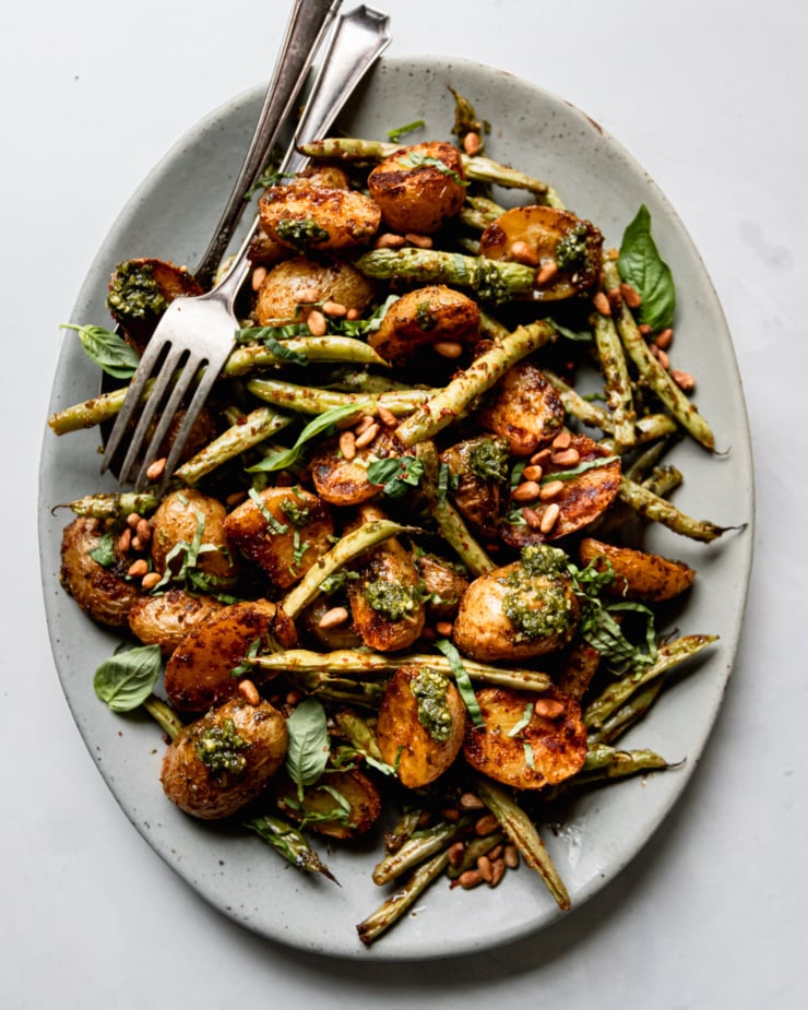 An overhead shot shows a platter of roasted green beans and potatoes dressed with a lemony pesto sauce. The veggies are garnished with fresh basil and toasted pine nuts. Serving utensils are perched on the platter as well.
