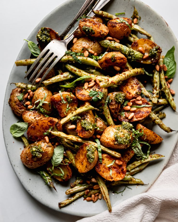 An up close, overhead shot shows a platter of roasted green beans and potatoes dressed with a lemony pesto sauce. The veggies are garnished with fresh basil and toasted pine nuts.