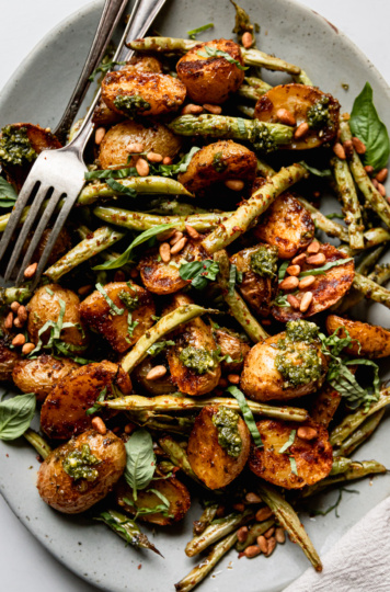 An up close, overhead shot shows a platter of roasted green beans and potatoes dressed with a lemony pesto sauce. The veggies are garnished with fresh basil and toasted pine nuts.
