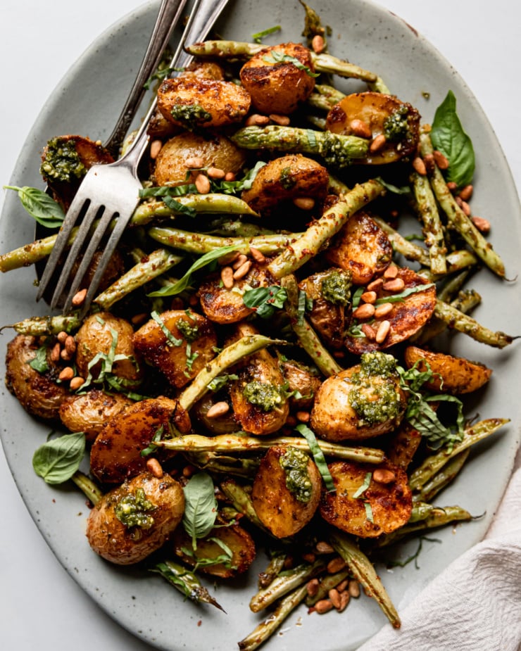An up close, overhead shot shows a platter of roasted green beans and potatoes dressed with a lemony pesto sauce. The veggies are garnished with fresh basil and toasted pine nuts.