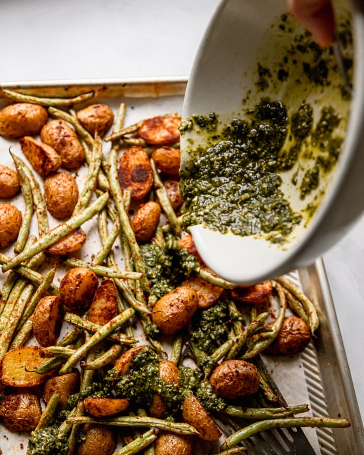 A 3/4 angle shot shows a pesto sauce being poured over a baking sheet of roasted vegetables.