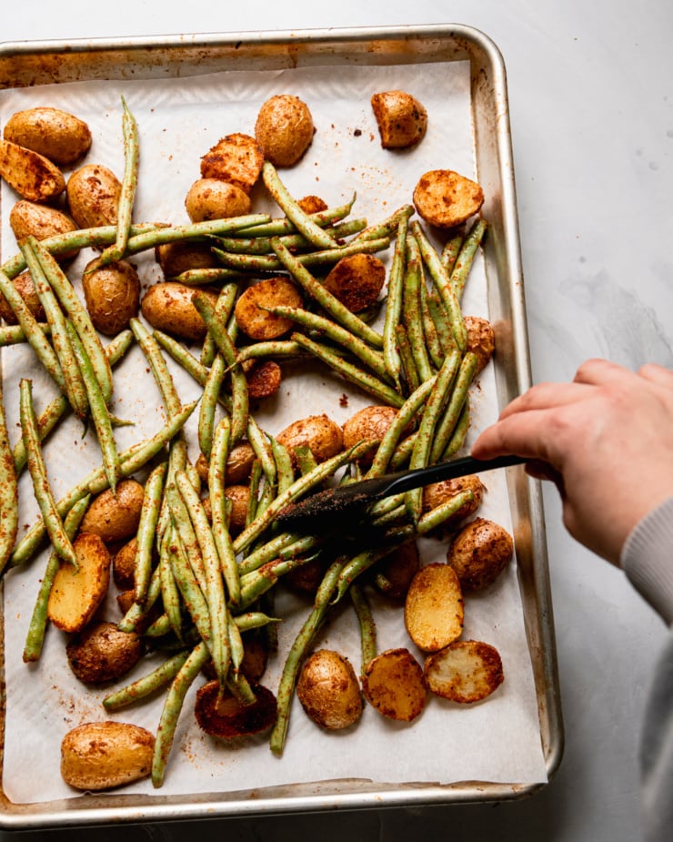 An overhead shot shows a hand using a spatula to distribute green beans on a baking sheet with roasted potatoes.