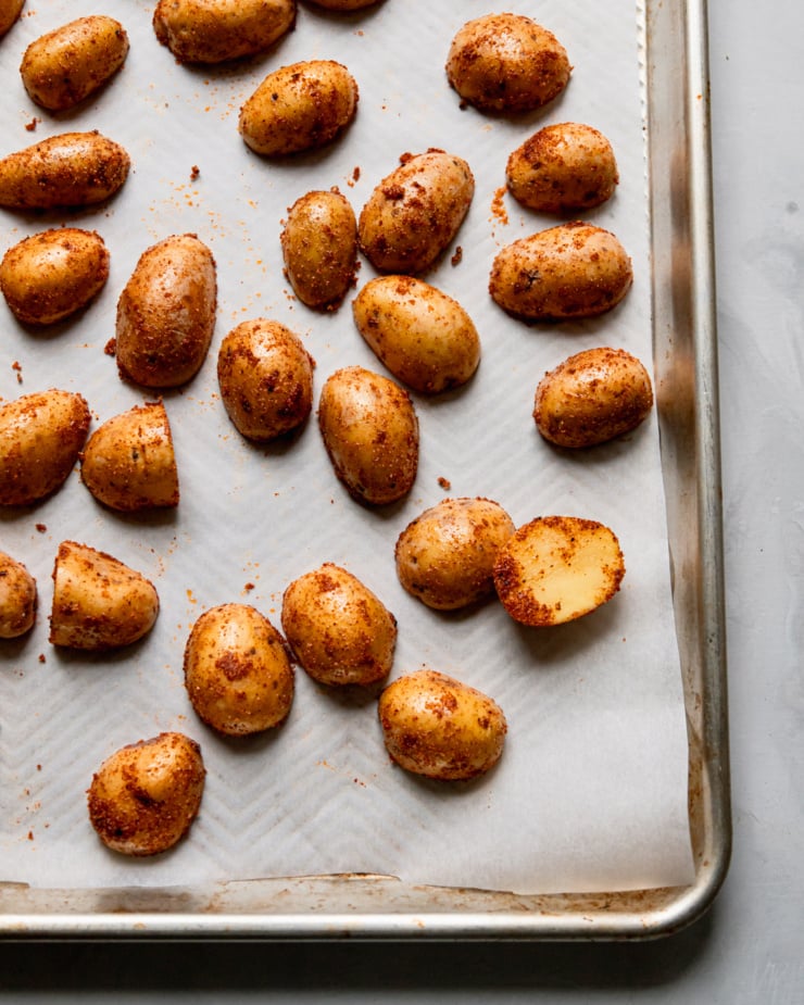 An overhead shot shows potatoes coated in oil and spices on a parchment-lined baking sheet.