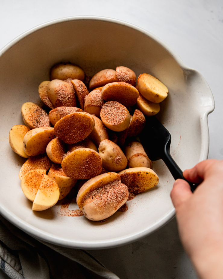 An overhead shot shows a hand using a spatula to toss halved baby Yukon gold potatoes in spices, salt, pepper, and oil in a bowl.