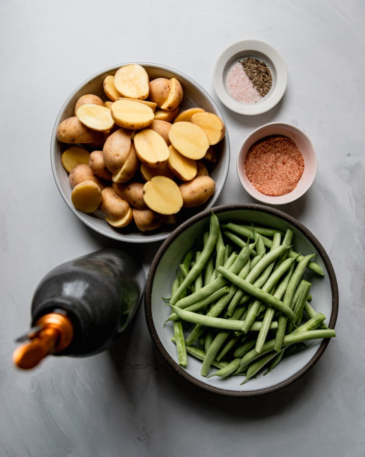 An overhead shot shows a bowl of halved baby potatoes, trimmed green beans, salt, pepper, a spice blend, and a bottle of olive oil.