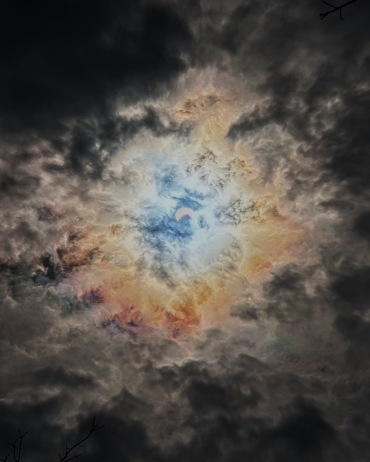 A photograph of the sky during the solar eclipse shows a crescent shaped glow from the sun surrounded by dark clouds.