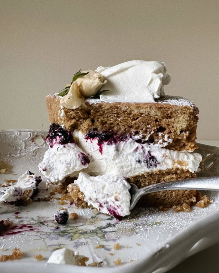 A head-on shot shows a slice of cake with a coconut cream and lavender blueberry compote layer. A white rose flower adorns the top of the slice.