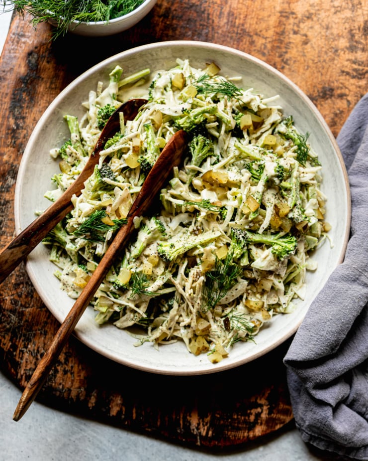 An overhead shot shows a bowl of creamy and crunchy dill pickle broccoli slaw with cabbage, broccoli, dill pickles, fresh dill, and a creamy dill pickle dressing. Wooden tongs are sticking out of the bowl.