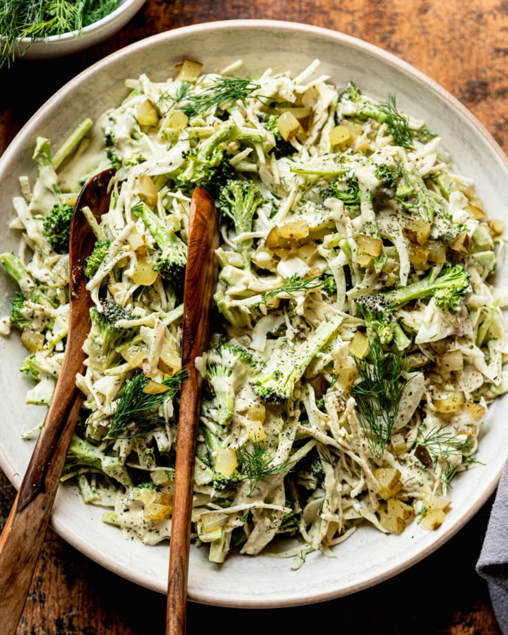An overhead shot shows a bowl of creamy and crunchy dill pickle broccoli slaw with cabbage, broccoli, dill pickles, fresh dill, and a creamy dill pickle dressing. Wooden tongs are sticking out of the bowl.