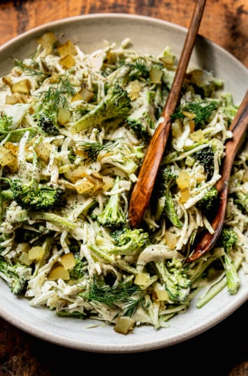 An overhead shot shows a bowl of creamy and crunchy dill pickle broccoli slaw with cabbage, broccoli, dill pickles, fresh dill, and a creamy dill pickle dressing. Wooden tongs are sticking out of the bowl.