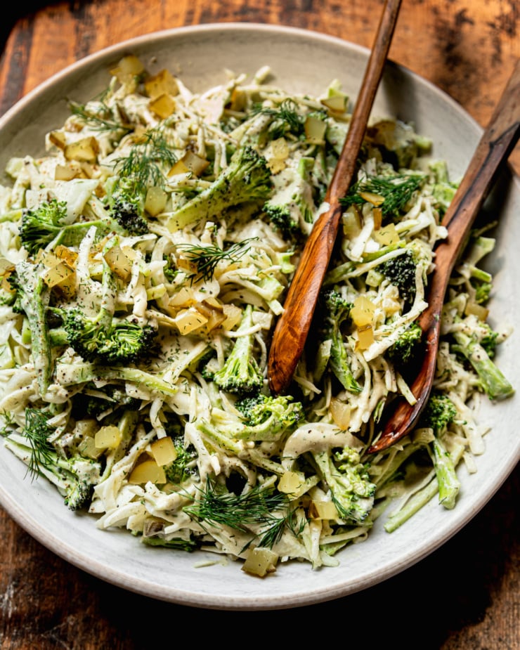 An overhead shot shows a bowl of creamy and crunchy dill pickle broccoli slaw with cabbage, broccoli, dill pickles, fresh dill, and a creamy dill pickle dressing. Wooden tongs are sticking out of the bowl.