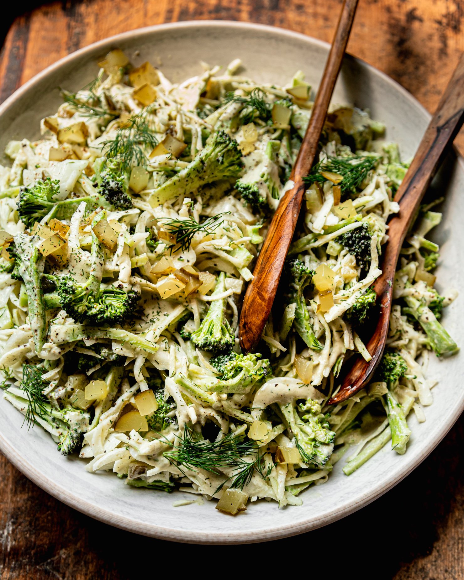 An overhead shot shows a bowl of creamy and crunchy dill pickle broccoli slaw with cabbage, broccoli, dill pickles, fresh dill, and a creamy dill pickle dressing. Wooden tongs are sticking out of the bowl.