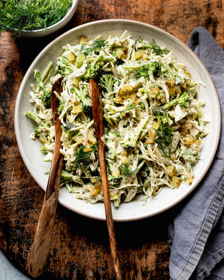 An overhead shot shows a bowl of creamy and crunchy dill pickle broccoli slaw with cabbage, broccoli, dill pickles, fresh dill, and a creamy dill pickle dressing. Wooden tongs are sticking out of the bowl.