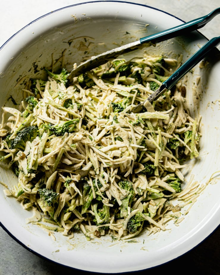 An overhead shot shows a creamy broccoli and cabbage slaw, all mixed up in an enamelware mixing bowl with tongs sticking out. The slaw appears creamy and well-dressed.