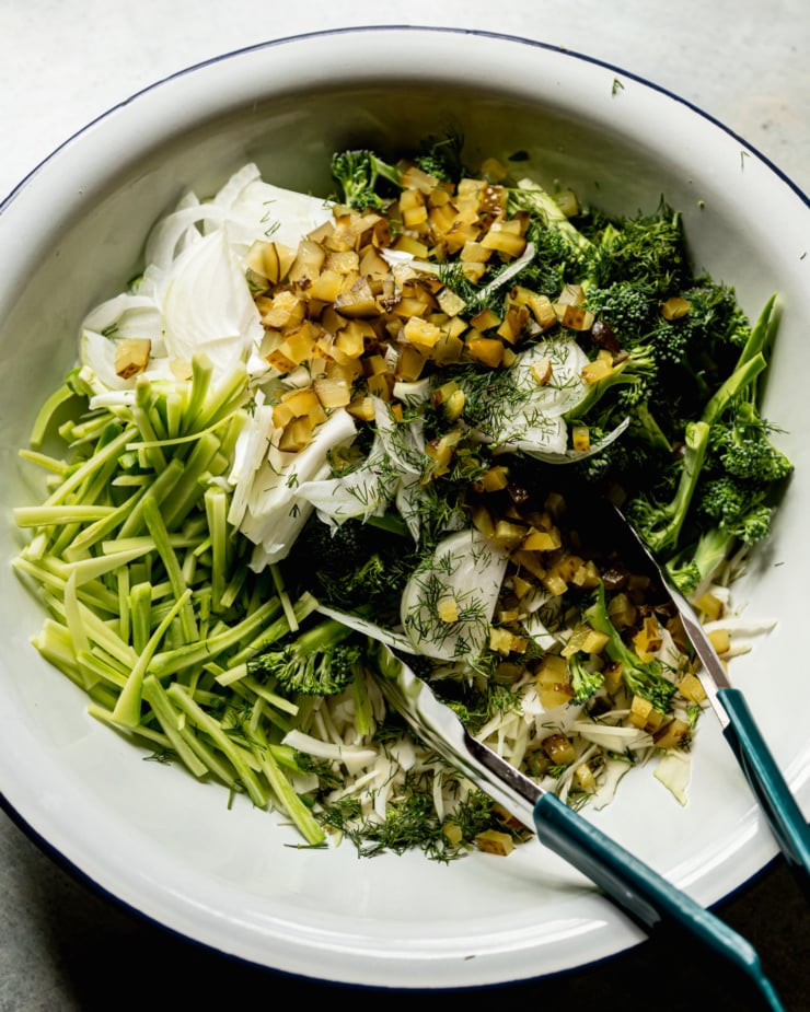 An overhead shot shows all of the prepped slaw ingredients in a large enamelware mixing bowl with tongs sticking out.