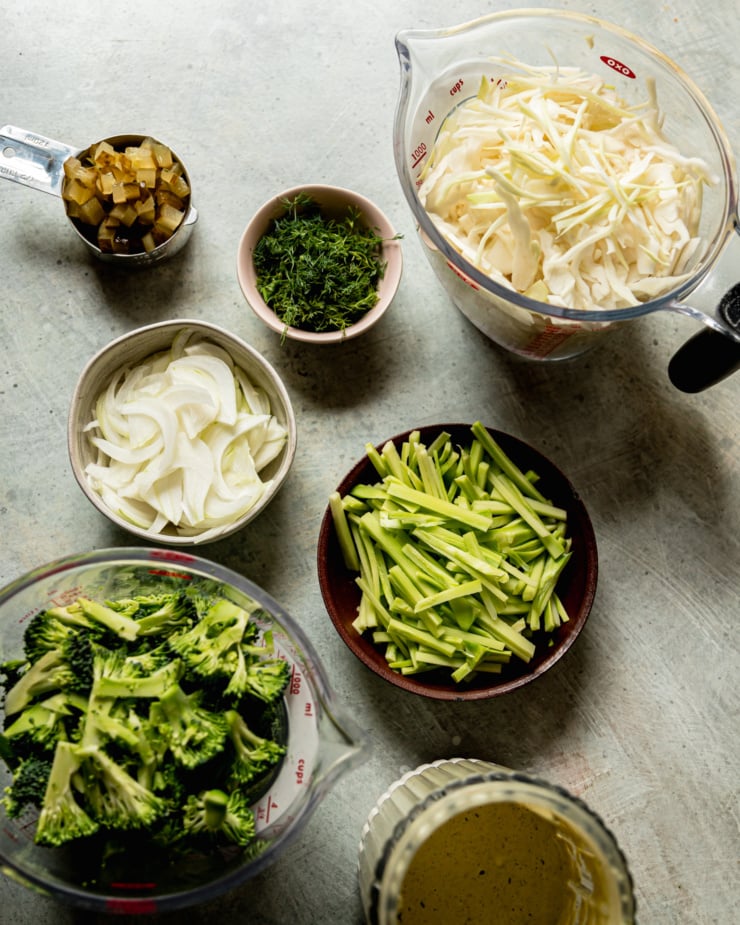 An overhead shot shows all of the prepped ingredients for a creamy dill pickle broccoli slaw: shredded cabbage, julienned broccoli stems, creamy dill pickle dressing, finely chopped broccoli florets, sliced sweet onion, chopped fresh dill, and chopped dill pickles.