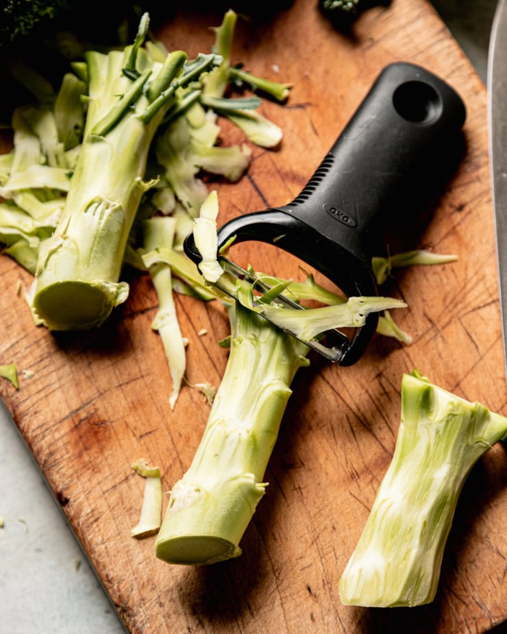 An overhead shot shows broccoli stalks having the tough outer layer peeled off with a vegetable peeler.