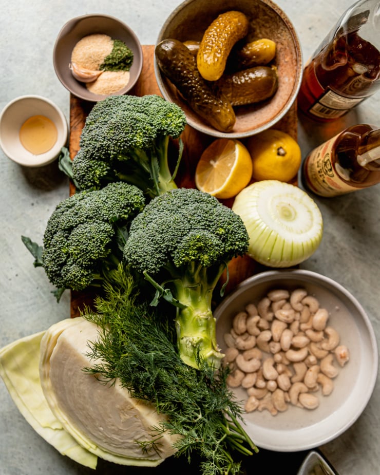 An overhead shot shows ingredients used in a dill pickle broccoli slaw recipe: pickles, white wine vinegar, worcestershire sauce, sweet onion, lemon, garlic, cashews, dill, cabbage, broccoli, agave nectar, spices, and garlic.