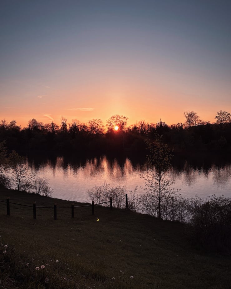 A head-on shot shows the sun rising above a tree line and river.