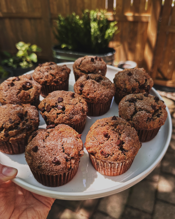 An overhead shot shows a hand holding a plate of banana chocolate chip muffins in dappled sunlight.