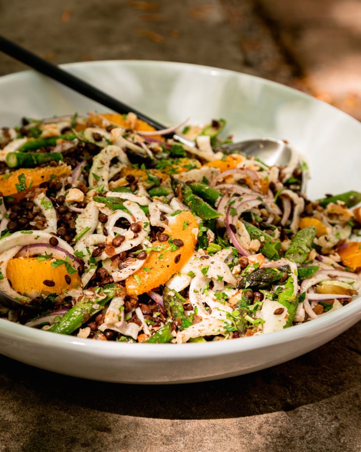 A head-on shot shows a bowl with a lentil salad and tongs sticking out. The salad features shaved fennel and red onion, sautéed diced asparagus, orange supremes, chopped parsley, walnuts, and a light vinaigrette.