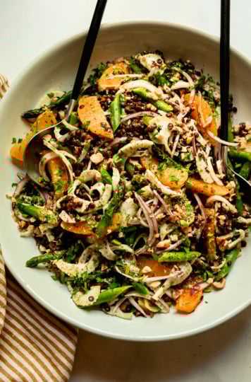 An overhead shot shows a french lentil salad with thinly sliced fennel and red onion, sautéed asparagus, orange supremes, chopped parsley and walnuts. Tongs are sticking out of the serving bowl.