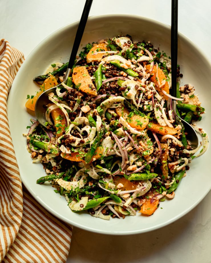 An overhead shot shows a french lentil salad with thinly sliced fennel and red onion, sautéed asparagus, orange supremes, chopped parsley and walnuts. Tongs are sticking out of the serving bowl.