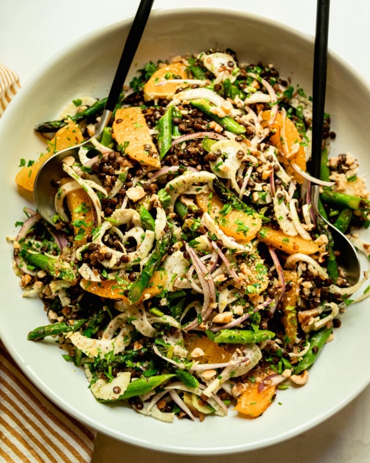 An overhead shot shows a french lentil salad with thinly sliced fennel and red onion, sautéed asparagus, orange supremes, chopped parsley and walnuts. Tongs are sticking out of the serving bowl.