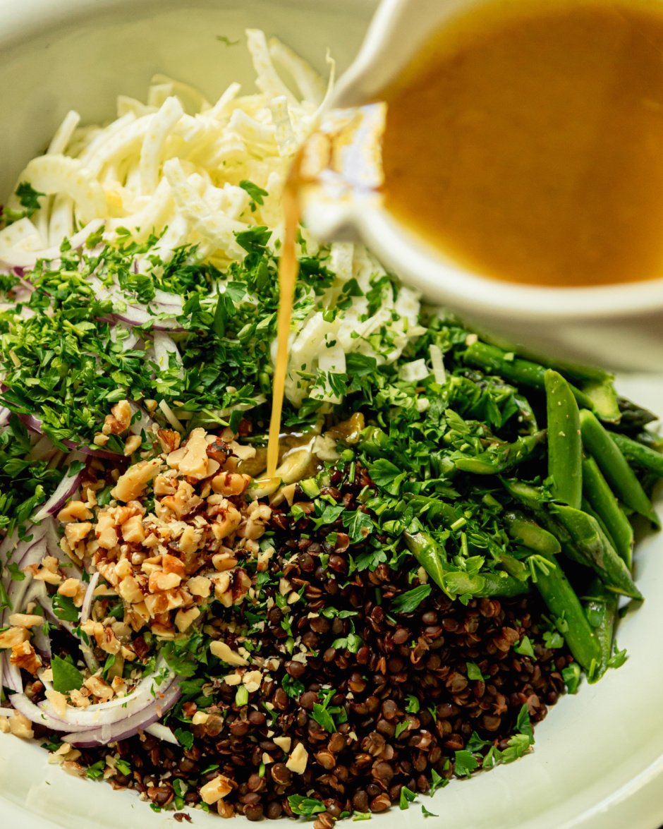 A slight 3/4 angle image shows a cinaigrette being poured over sliced fennel, sautéed asparagus, chopped parsley, chopped walnuts, cooked French lentils, and sliced red onion in a large bowl.
