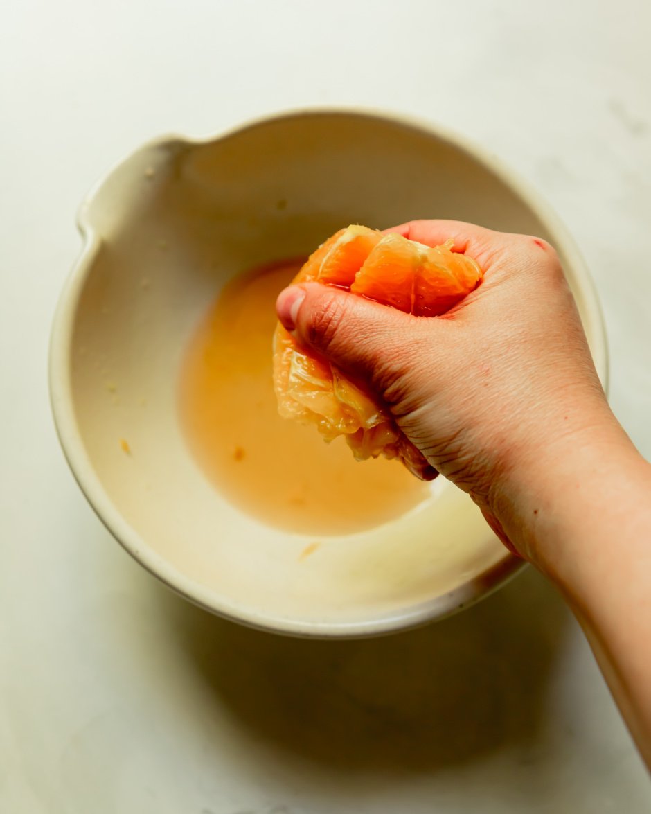An overhead shot shows a hand squeezing a portion of an orange to get the juice in a bowl below.