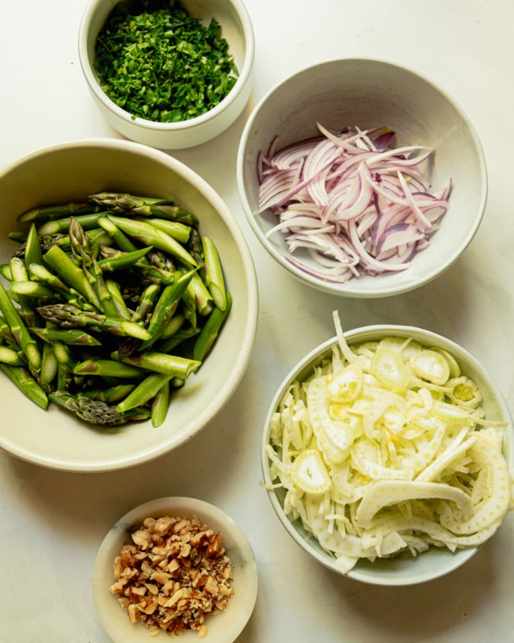 An overhead shot shows prepped ingredients in bowls: chopped parsley, sliced red onion, sliced fennel, chopped walnuts, chopped asparagus.