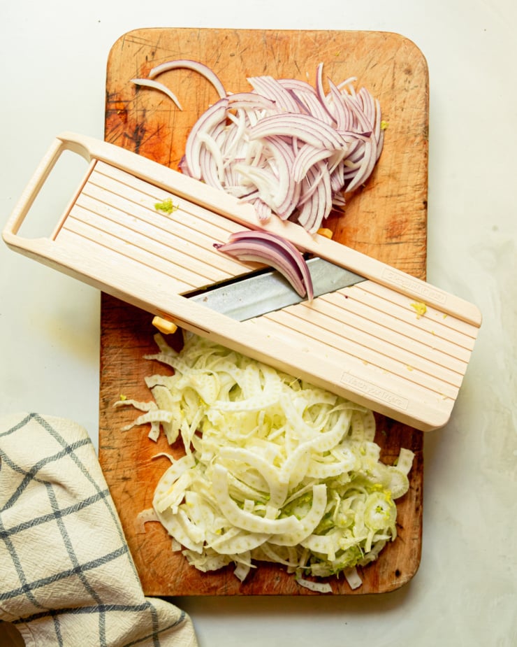 An overhead shot shows a wooden cutting board filled with thin red onion and fennel slices. A mandolin slicer is perched in the middle.