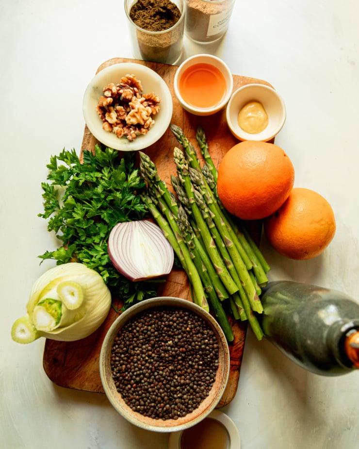 An overhead shot shows ingredients for a salad: spices, apple cider vinegar, dijon mustard, oranges, asparagus, olive oil, french lentils, red onion, parsley, fennel, and walnuts.