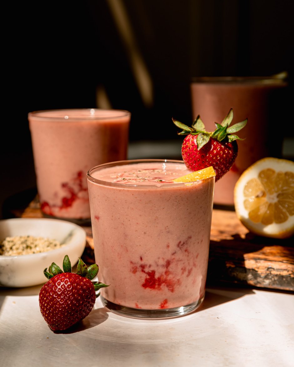 A head-on shot in direct sunlight shows a strawberry lemonade smoothie garnished with a whole strawberry and a little lemon slice. There's a couple more glasses of the smoothie in the background.