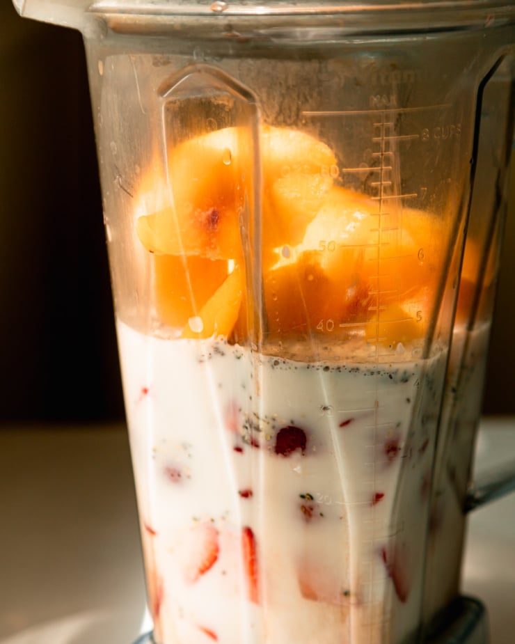 A head-on shot shows ingredients like non-dairy milk, frozen peaches, and strawberries in a blender, about to be blended up.