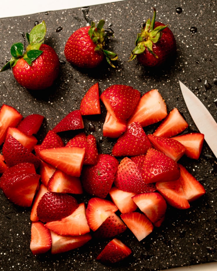 An overhead shot shows strawberries being chopped on a cutting board with a paring knife.