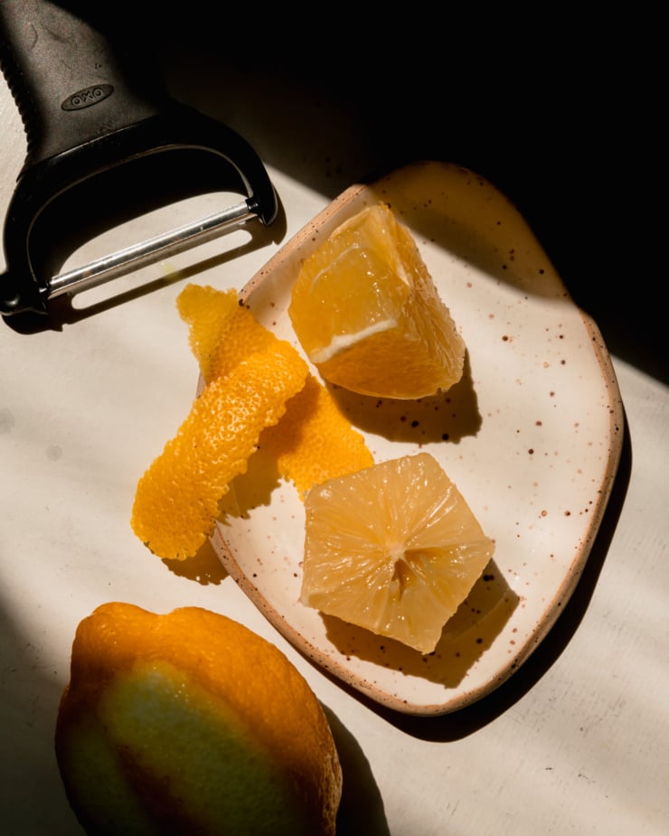 An overhead shot in direct sunlight shows a lemon that has been peeled and halved and two strips of lemon peel.