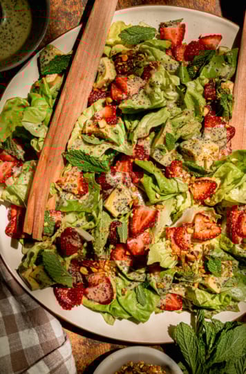 An overhead shows a plated strawberry salad with torn butter lettuce, chopped pistachios, diced avocado, mint leaves, and lemony mint poppy seed dressing. Wooden salad tongs are sticking out of the salad.
