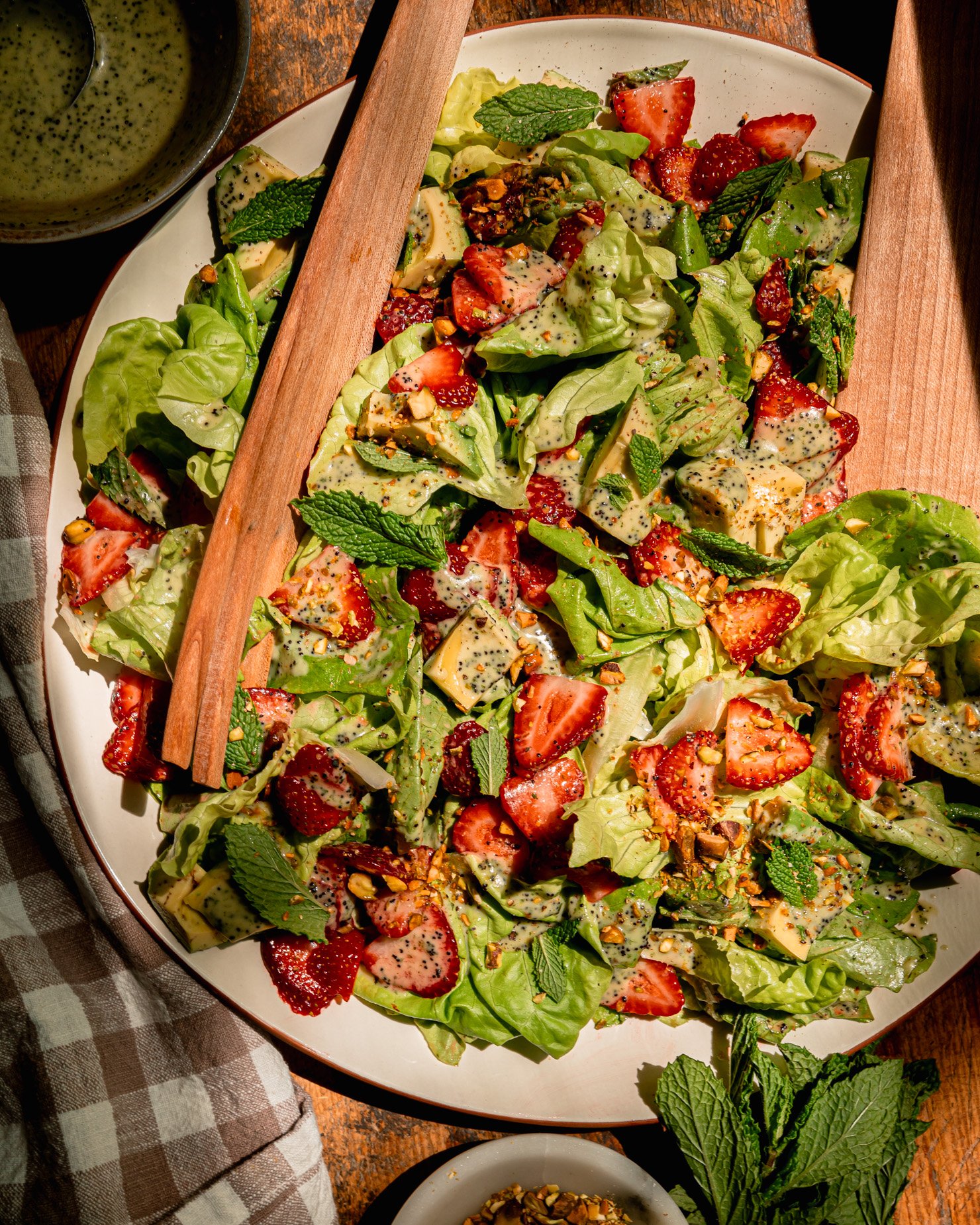 An overhead shows a plated strawberry salad with torn butter lettuce, chopped pistachios, diced avocado, mint leaves, and lemony mint poppy seed dressing. Wooden salad tongs are sticking out of the salad.