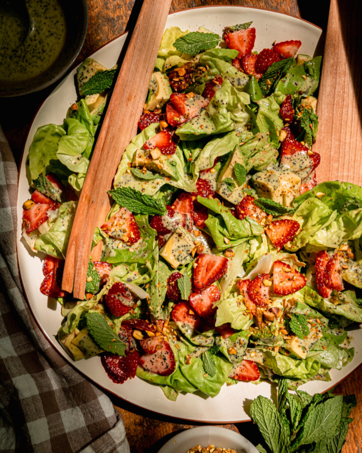 An overhead shows a plated strawberry salad with torn butter lettuce, chopped pistachios, diced avocado, mint leaves, and lemony mint poppy seed dressing. Wooden salad tongs are sticking out of the salad.