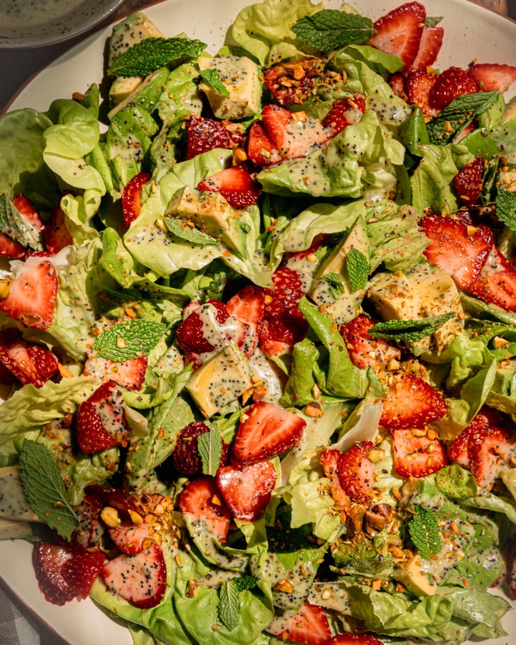 An overhead shot shows a plated strawberry salad with torn butter lettuce, chopped pistachios, diced avocado, mint leaves, and lemony mint poppy seed dressing.