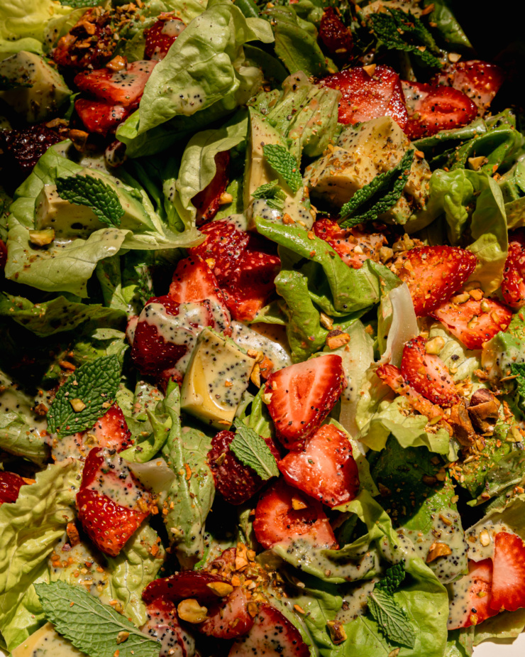 AN up close overhead shot shows a strawberry salad with torn butter lettuce, chopped pistachios, diced avocado, mint leaves, and lemony mint poppy seed dressing. The photo is taken in direct sunlight.