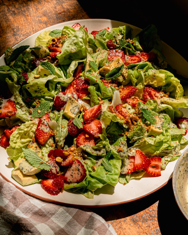 A 3/4 angle image shows a plated strawberry salad with torn butter lettuce, chopped pistachios, diced avocado, mint leaves, and lemony mint poppy seed dressing. A checked linen napkin is nearby.
