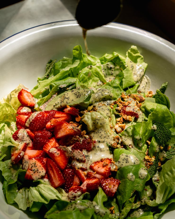 A 3/4 angle shot shows lemony mint poppy seed dressing being poured over torn butter lettuce, sliced strawberries, mint leaves, and chopped pistachios in direct sunlight.