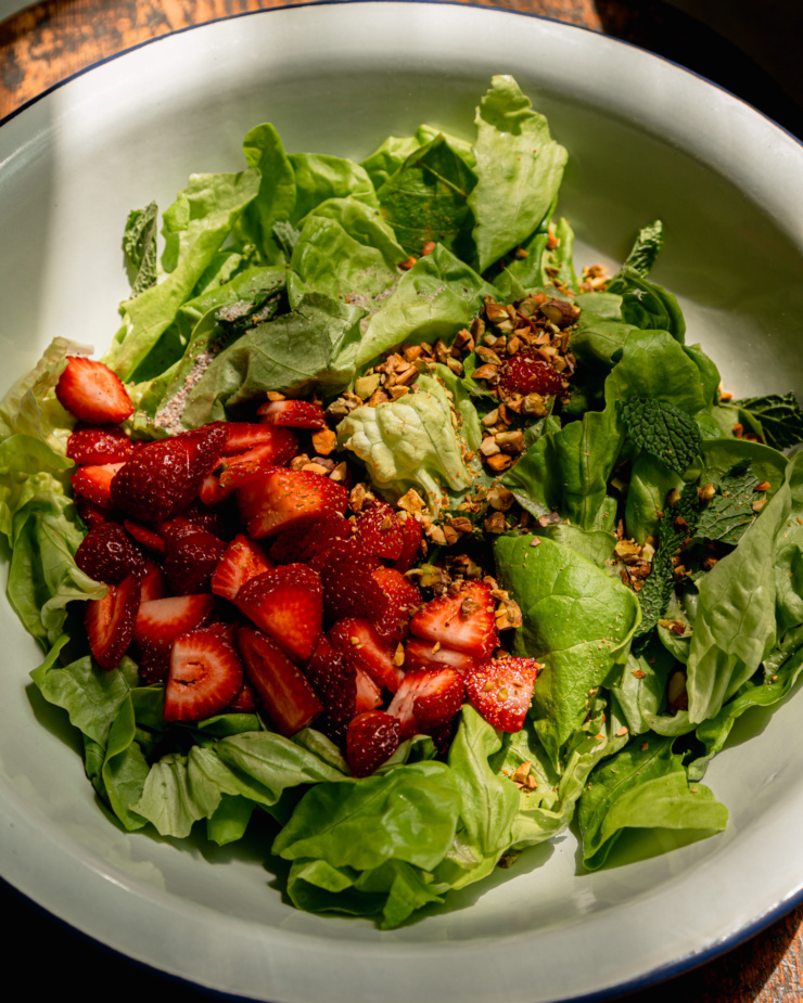 A 3/4 angle shot shows the following in a large bowl: torn butter bettuce, mint leaves, sliced strawberries, and chopped pistachios.