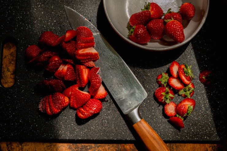An overheard shot shows strawberries being hulled and sliced on a cutting board.