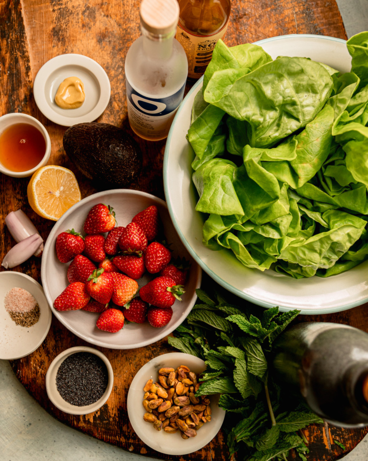 An overhead shot shows ingredients for a strawberry salad with butter lettuce, lemon mint poppy seed dressing, pistachios, and avocado.
