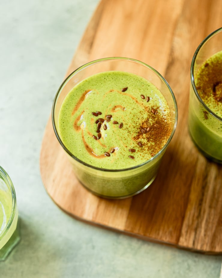 An overhead shot shows a glass filled with a peanut butter green smoothie. The top is dusted with cinnamon and garnished with swoops of peanut butter and flax seeds.