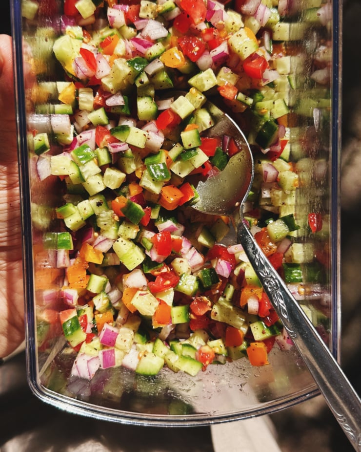An overhead shot shows finely diced cubes of cucumber, tomato, and red onion seasoned with sumac, lemon, salt, pepper, and olive oil. A spoon is sticking out of the rectangular container.