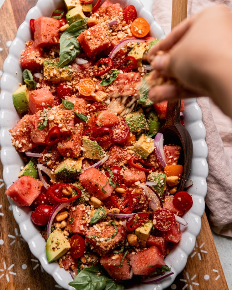 An overhead shot shows a hand sprinkling finely crushed salted corn nuts over a watermelon salad in an oval-shaped platter.